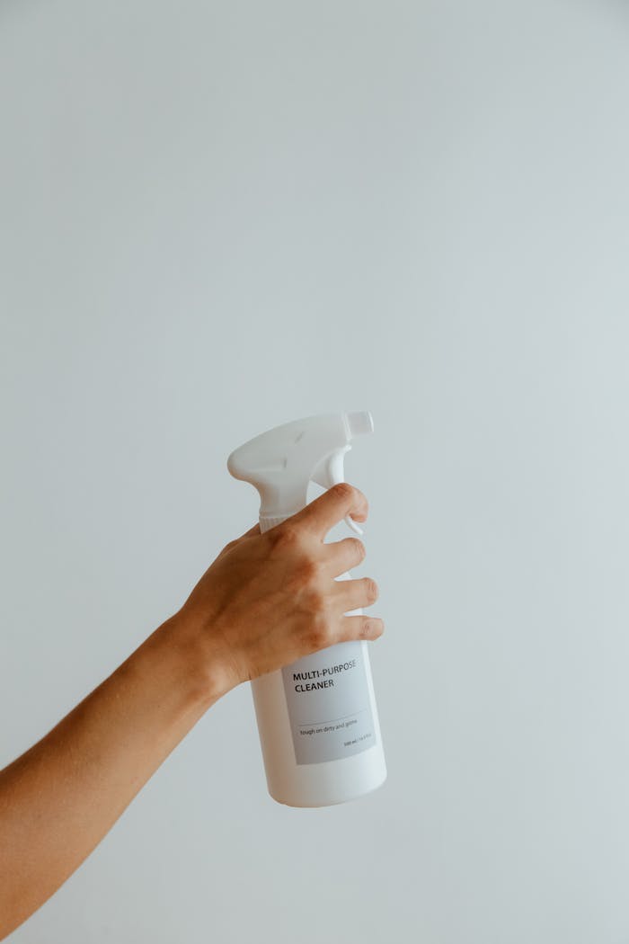 services-02 Close-up of a hand holding a white multipurpose cleaner spray bottle against a minimalistic background.